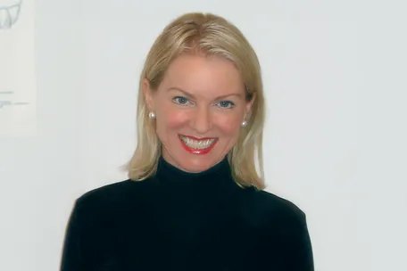 Porträt einer lächelnden Frau mit hellblonden Haaren und schwarzem Rollkragenpullover vor hellem Hintergrund.