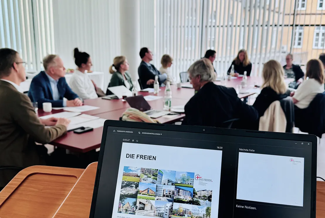 Gruppe von Menschen in einem hellen Raum bei einer Besprechung. Im Vordergrund ein Laptop mit einer Präsentation zu „Die Freien“ und Fotografien der verschiedenen Kliniken.
