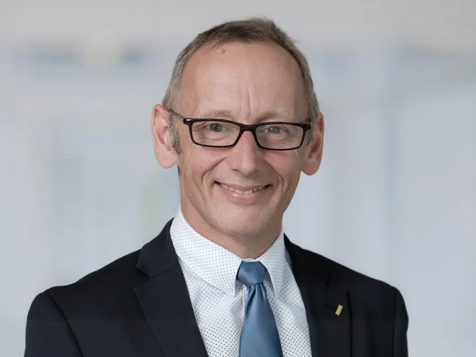 Porträt eines lächelnden Mannes mit Brille, schwarzem Anzug und hellblauer Krawatte vor unscharfem Hintergrund.