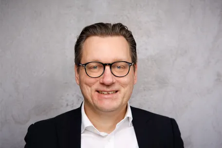 Porträt eines lächelnden Mannes mit Brille, schwarzem Sakko und weißem Hemd vor einem grauen Hintergrund.