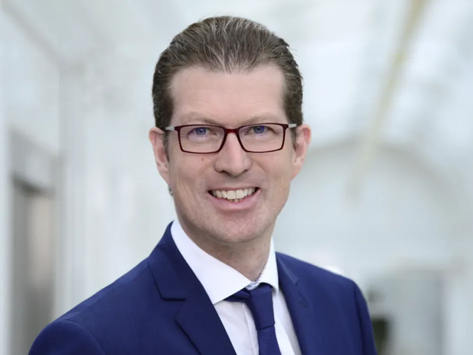 Porträt eines lächelnden Mannes mit Brille und dunkelblauem Anzug vor einem hellen, modernen Hintergrund.